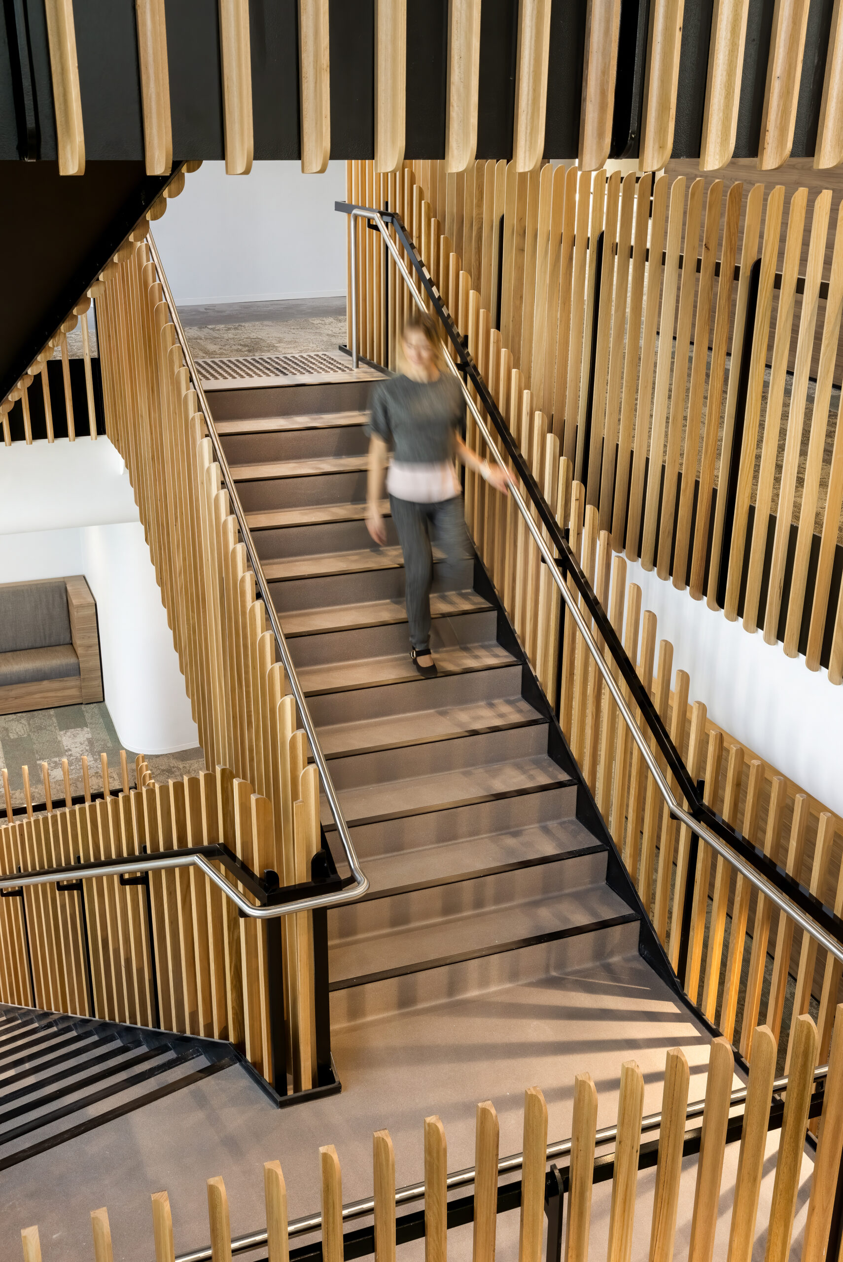 Modern staircase with warm wooden slats, sleek black railings and stair nosings, and a blurred person descending. Seating area visible on the left.