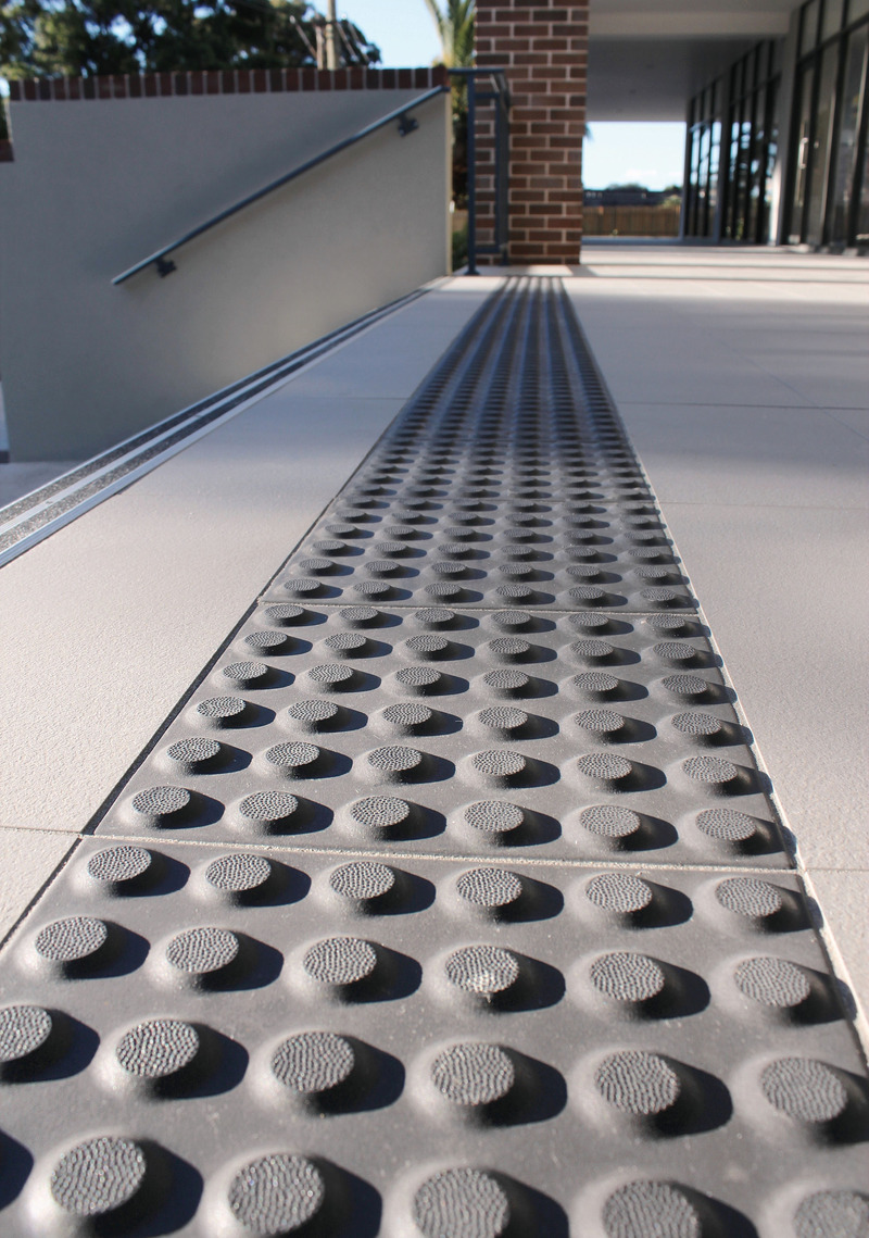 Close-up of a charcoal ceramic tactiles paving strip on a concrete walkway. It leads towards a brick wall and glass-fronted building.