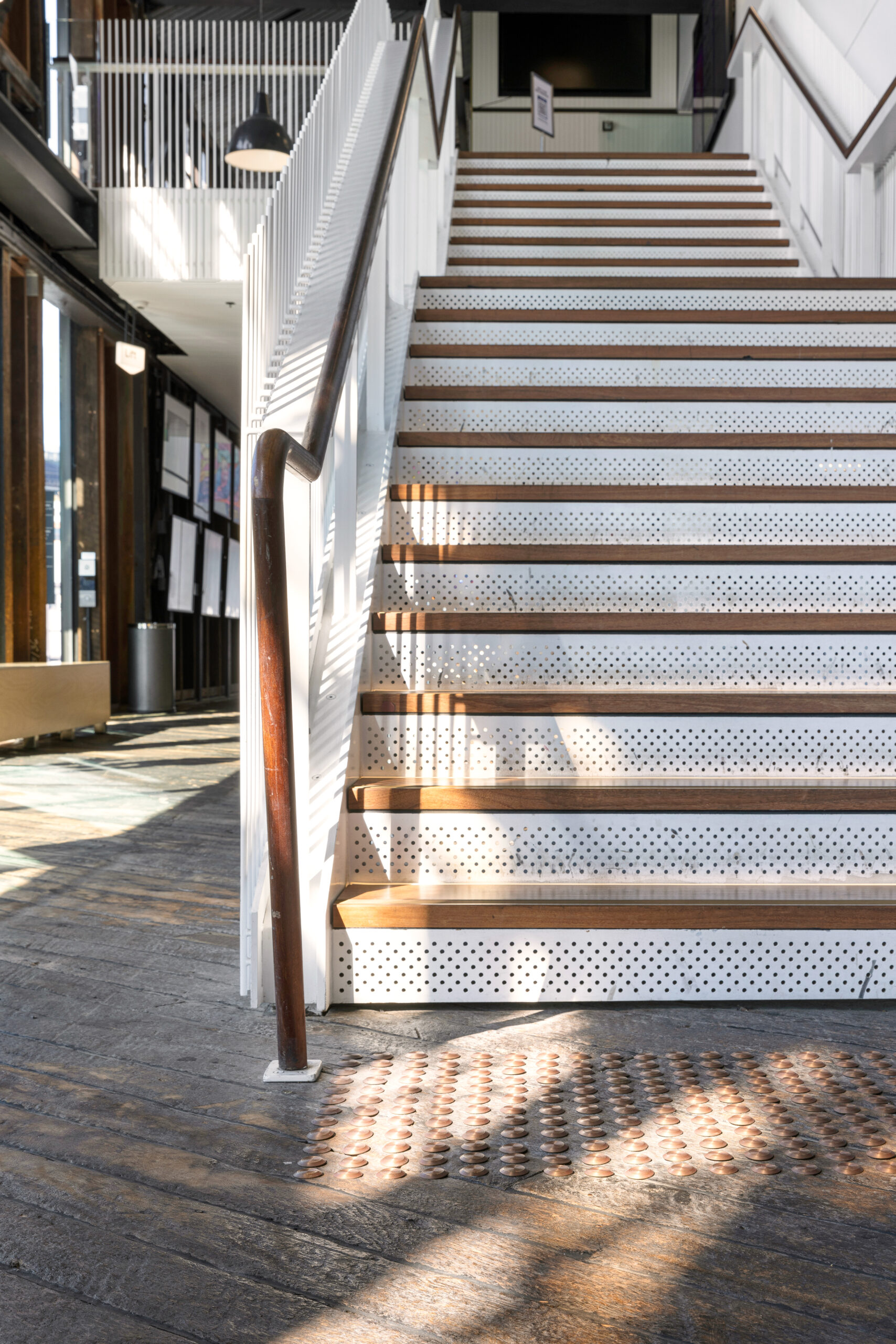 Sunlit staircase with solid brass stair nosings and tactile indicators in a modern building. Natural light casts patterns on the steps and floor.