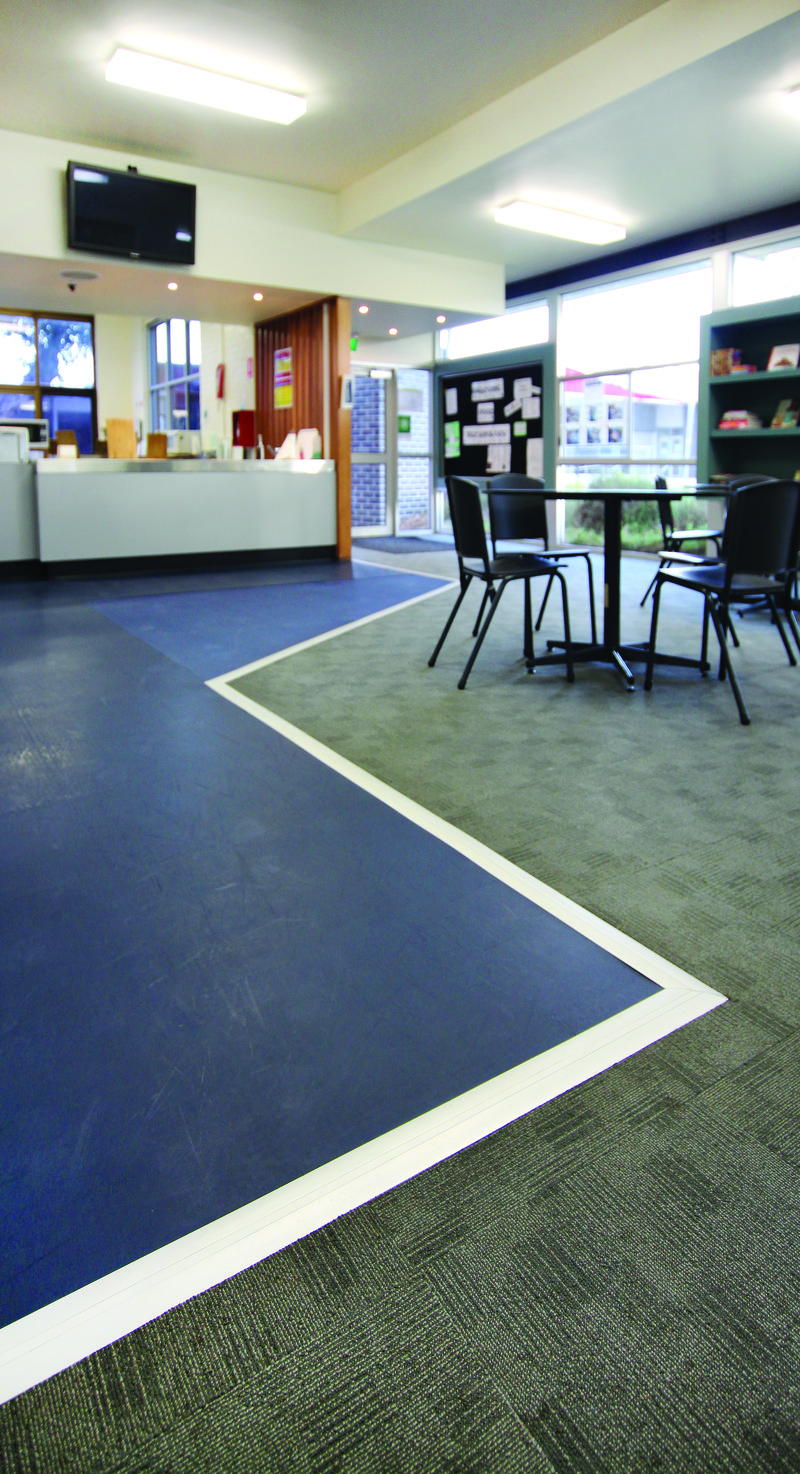 A modern office space with blue flooring and gray carpeted areas, separated by floor trim covers. There is an empty round table with chairs and natural light from large windows.
