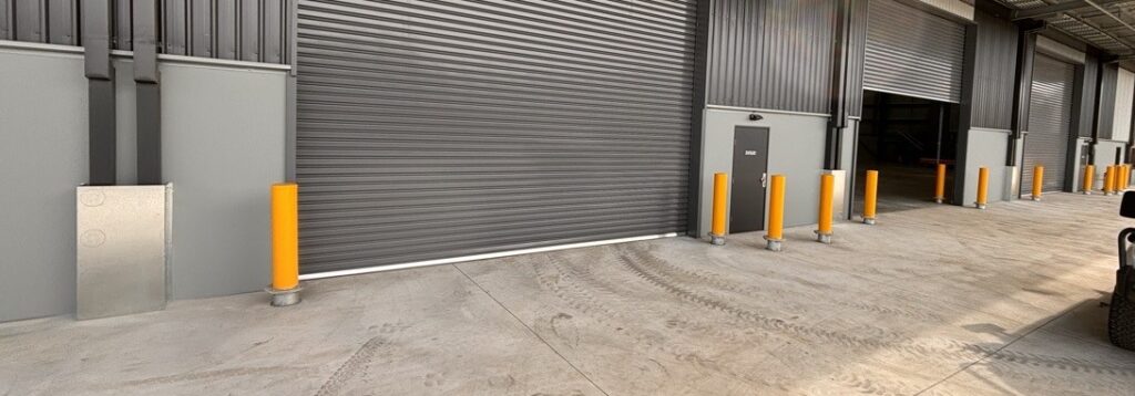 Yellow powder-coated breakaway bollards mid-installation next to roller shutters in an industrial property.