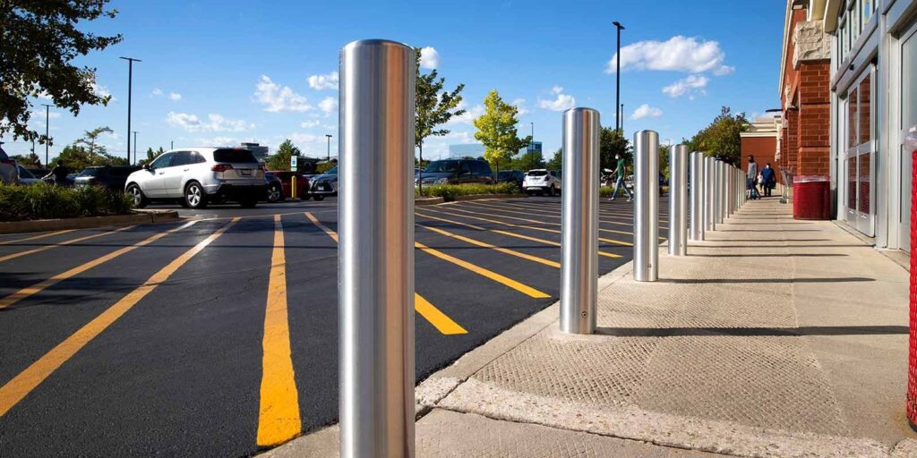 In-ground bollards installed on the pedestrian pavement between a car park and a commercial building.