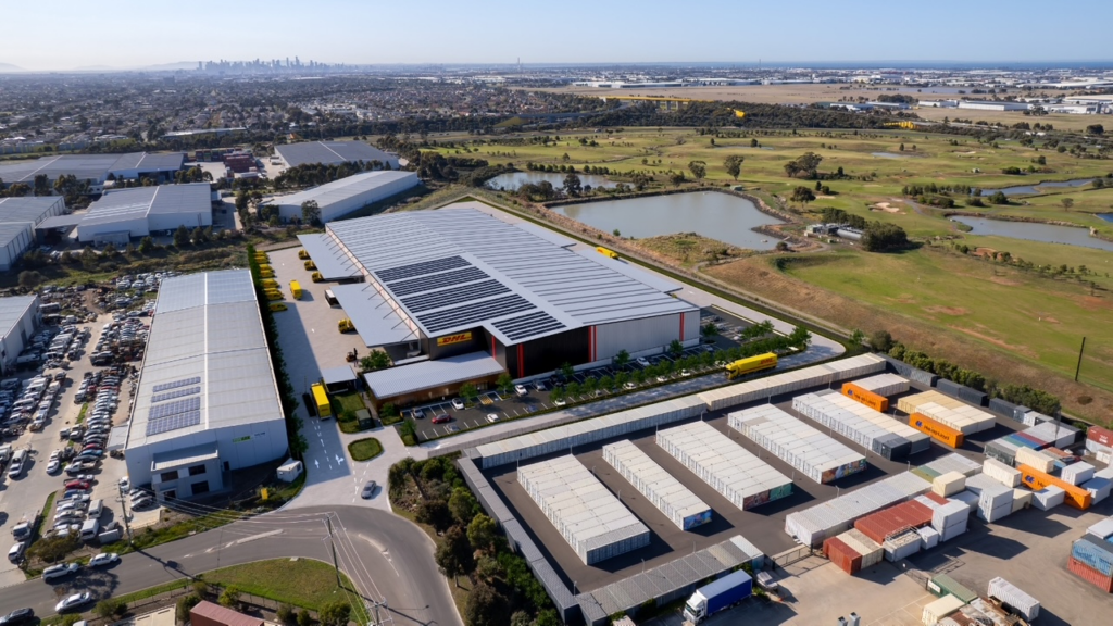 Aerial view of a large industrial warehouse complex with solar panels on the roof, surrounded by trucks and green fields. 