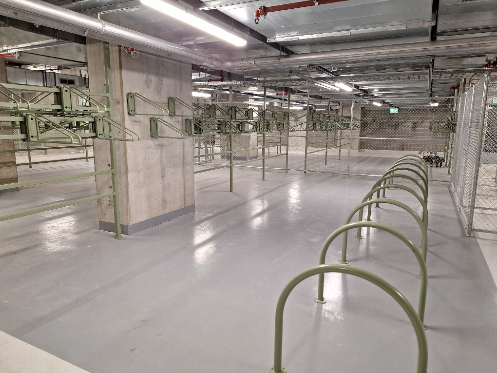 Spacious indoor bike storage with green bike hoops and vertical stands, under fluorescent lighting.